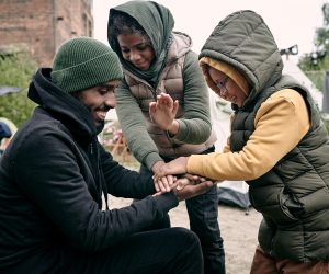Positive young Black man in hat playing with kids against abandoned building and hanging clothes on rope in refugee camp